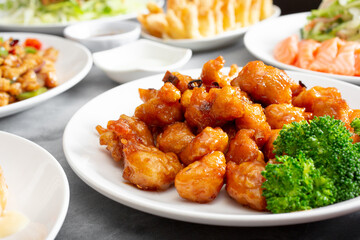 A view of a table of various Chinese entrees, featuring a plate of orange chicken.
