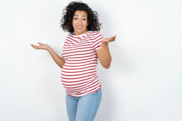 Clueless young pregnant woman wearing striped t-shirt over white background shrugs shoulders with hesitation, faces doubtful situation, spreads palms, Hard decision