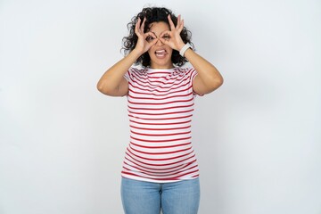 young pregnant woman wearing striped t-shirt over white background doing ok gesture like binoculars sticking tongue out, eyes looking through fingers. Crazy expression.