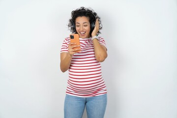 Happy young pregnant woman wearing striped t-shirt over white background feels good while focused in screen of smartphone. People, technology, lifestyle