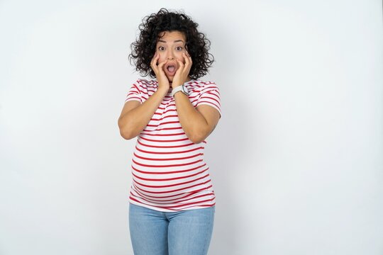 Stupefied young pregnant woman wearing striped t-shirt over white background expresses excitement and thrill, keeps jaw dropped, hands on cheeks, has eyes popped out