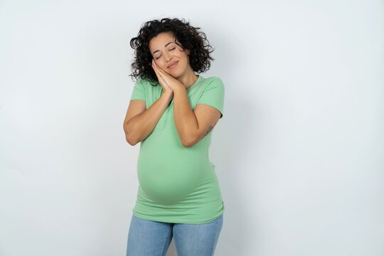 young pregnant woman wearing green t-shirt over white background leans on pressed palms closes eyes and has pleasant smile dreams about something