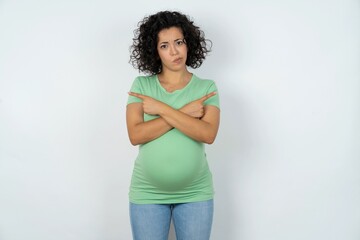 Serious young pregnant woman wearing green t-shirt over white background crosses hands and points...