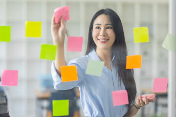 Smiling business Asian people thinking and use sticky notes on glass wall in office with her colleague in background.
