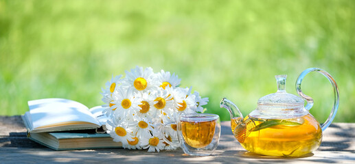 Obraz na płótnie Canvas Chamomile flowers, books, glass teapot and cup with herbal tea on table close up, natural abstract green background. summer season. relax time. useful calming tea. tea party in garden.