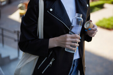 Thirsty young woman drinking mineral water from a glass bottle. Responsible female person using a reusable water bottle and wearing a cotton tote bag on shoulder