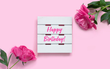 Bouquet of beautiful pink peonies with gift boxes in paper wrapping. White wooden board with text Happy Birthday!
