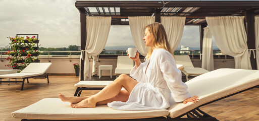 Woman in white bathrobe lying on sofa and relaxing with orange cocktail at the roof. - obrazy, fototapety, plakaty