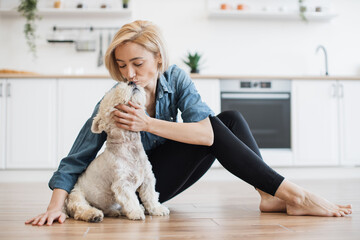Friendly caucasian female kissing white terrier's nose while resting on hard surface on kitchen...