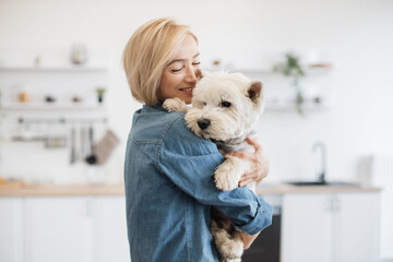 Beautiful woman in jeans shirt holding medium-sized dog on shoulder while standing in light room interior. Emotional female keeper finding joy in everyday interaction with furry friend indoors.