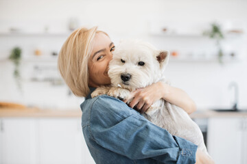 Portrait of smiling blonde lady with West Highland White Terrier in arms posing on background of...