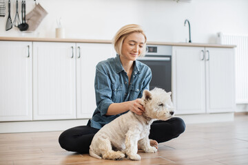 Happy caucasian female adult in casual clothes rubbing ears of small terrier while sitting cross-legged in kitchen. Smiling slim woman making her adorable furry friend feel relaxed during day indoors.