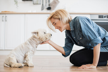 Vivid short-haired lady in casual clothes fondling West Highland White Terrier while sitting on bent knees indoors. Cheerful female pet lover keeping calm dog entertained during break from workout.