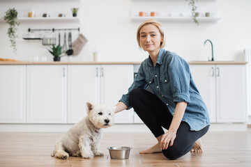 Portrait of beautiful relaxed woman and well-trained white terrier posing in modern kitchen of spacious house. Confident pet owner finishing brief indoor training session with homemade treats.