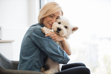 Close up portrait of charming blonde woman cuddling adorable Westie while seating in armchair in apartment. Affectionate calm lady in cozy clothes strengthening friendship bond with canine buddy.