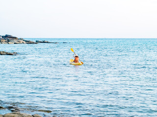 Back of Asian woman in life jacket and cap kayaking on yellow kayak boat with using paddle on the sea. Happy female having fun activity on seascape view background, Holiday trip vacation.