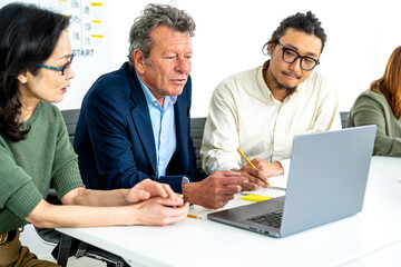 Multiracial group working at meeting table in a white office, focuns on senior business man looking at laptop