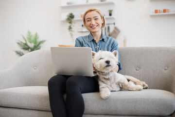 Smiling adult woman and sleepy purebred Westie posing with computer on laps in comfortable room with soft couch. Charming short-haired blonde scrolling digital photos from last birthday party.