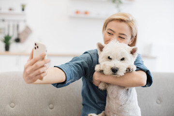 Close up view of vivid blonde lady in denim shirt looking at front camera of mobile while hugging smart terrier indoors. Lovely woman getting self-taken picture with furry friend on room background.
