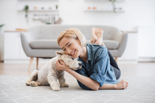 Pretty smiling woman in casual wear snuggling to cute pet's head while testing comfort of wooden floor with carpet on it. Joyful healthy lady appreciating affection and support of her canine friend.