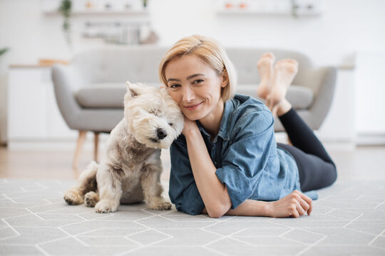 Full length portrait of lovely smiling woman in denim shirt caressing adorable little Westie on kitchen floor of apartment. Cheerful pet keeper posing with good-natured small animal in home interior.