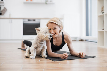 Portrait of happy blonde woman snuggling to adorable little dog while holding Forearm Plank Pose at home. Beautiful slim lady leading healthy lifestyle doing yoga whilst caring for furry friend.