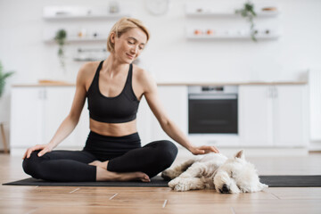Charming blonde female in black top and yoga pants stroking gently sleepy dog while resting with legs crosses on mat. Caring fit yogini taking break in yoga session for caressing pet at home.