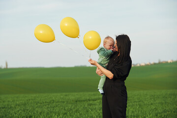 With yellow colored balloons. Mother with her little baby son is outdoors on the agricultural field