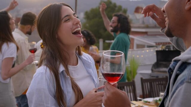 Joyful multi-ethnic couple dancing and drinking red wine celebrating rooftop party at sunset. Excited young friends having fun in background. People outdoors on summer vacation. 