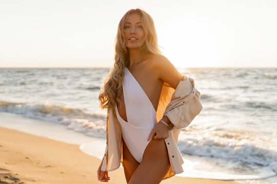 Seductive  blond woman in sexy swimwear posing on the beach in sunset light. Prtfect wavy hairs, tan skim body. Summer tropical mood.