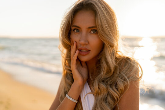  Close up portrait of beautiful blond woman in sexy swimwear posing on the beach in sunset light. Prtfect wavy hairs, tan skim body. Summer tropical mood.