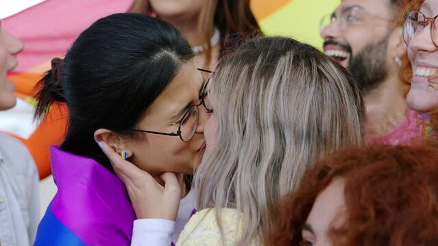 Happy young group of diverse people celebrating gay pride festival day together. Lesbian couple kissing showing love surrounded by friends. LGBT youth community concept.