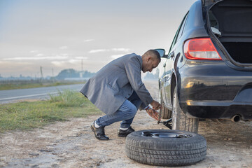 Latino man on suit changes a flat tire on his car at the side of the road with copy space.