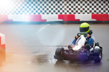 Young brave driver in karting dressed in a protective helmet participates in a karting race on a wet track. Little boy rides a go-kart on the track on a children's circuit - obrazy, fototapety, plakaty