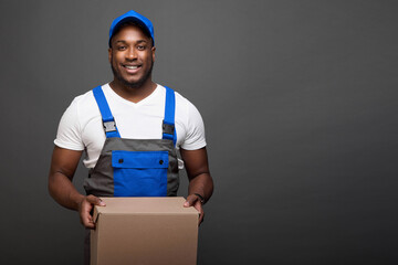 Cheerful dark-skinned deliveryman easily holds a heavy cardboard box in his hands. Black guy in...