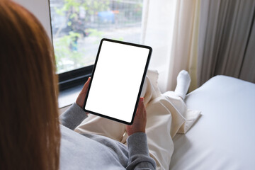 Mockup image of a woman holding digital tablet with blank desktop white screen while sitting on a bed