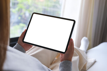 Mockup image of a woman holding digital tablet with blank desktop white screen while sitting on a bed