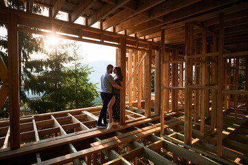 Man and woman examining their future wooden frame dwelling nestled in the mountains near forest. Youthful couple at construction site in early morning. Concept of contemporary ecological construction.