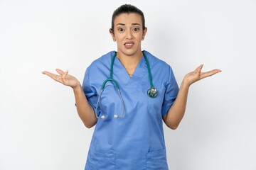 Clueless Beautiful doctor woman standing over white studio background shrugs shoulders with...