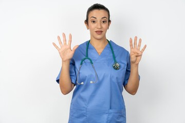 Beautiful doctor woman standing over white studio background showing and pointing up with fingers number nine while smiling confident and happy.