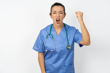 Beautiful doctor woman standing over white studio background angry and mad raising fist frustrated...