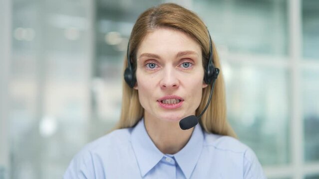 Webcam view. Woman in wireless headset customer service operator talking on video call while sitting in office. Agent of the call center advises the client, answering questions while looking in camera