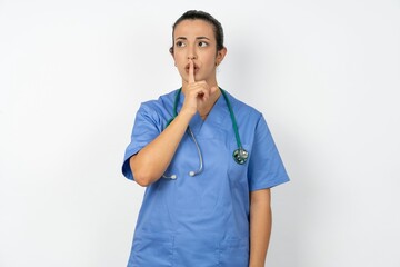 Beautiful doctor woman standing over white studio background silence gesture keeps index finger to...