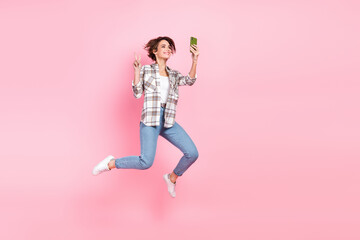 Full size portrait of pretty carefree girl jumping hold smart phone make selfie demonstrate v-sign isolated on pink color background