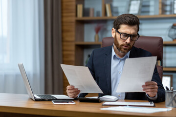 Serious and focused financier accountant on paper work inside office, mature man using calculator and laptop for calculating reports and summarizing accounts, businessman at work .