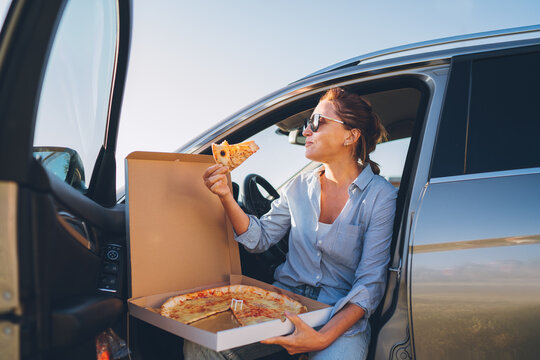 Midle-aged woman eating just cooked italian pizza sitting on driver car seat during meal break and enjoying sunset light. Auto traveling, fast food eating or car jorney lunch break concept image.