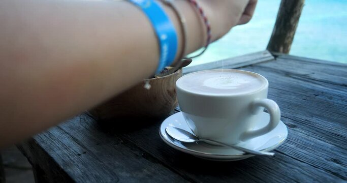 Handheld slow motion shot of a fresh caffee latte with a delicious bowl on the side while sugar is added with the blue sea with calm waves in the background
