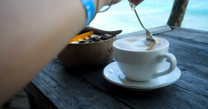 Handheld slow motion shot of a fresh caffee latte with a delicious bowl on the side while it is stirred with the blue sea with calm waves in the background