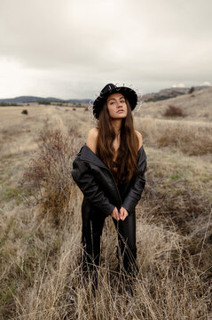 A beautiful model girl with wavy hair and bare shoulders in black leather jacket and a leather hat stands in a autumn field on a cloudy day among the yellow grass
