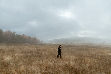 Misty autumn landscape with a girl in black look and silhouettes of horses in the distance. Landscape of overcast autumn with yellow grass, forest and a silhouette far away girl with in the mist